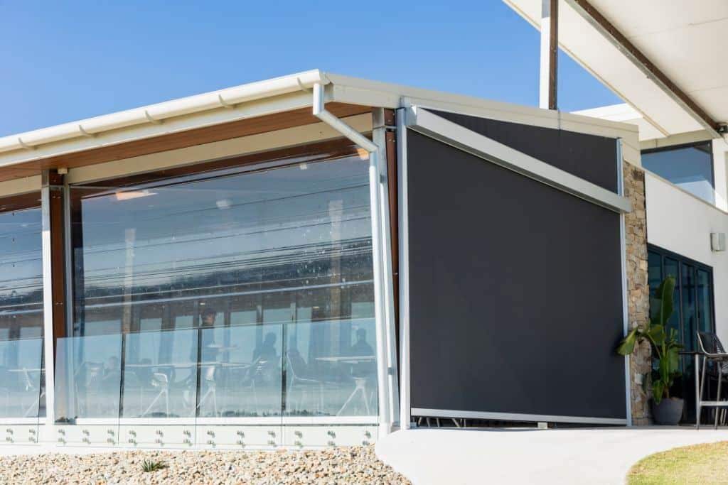AD 4nXdvhoawzZTbg5mri6MUbQUNWSOrJnfFPMGPfiDnILxKaJXO5adMfFb7G40BBfgnWpgmiC lNMLRfqMsIs5nZ btK HEj6jie iC5 Modern café with glass walls and a large black outdoor retractable blind, providing sun protection and wind resistance.