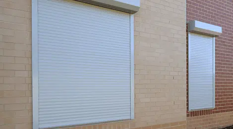 A brick building with closed white roller shutters, offering security, insulation, and light control for residential windows.